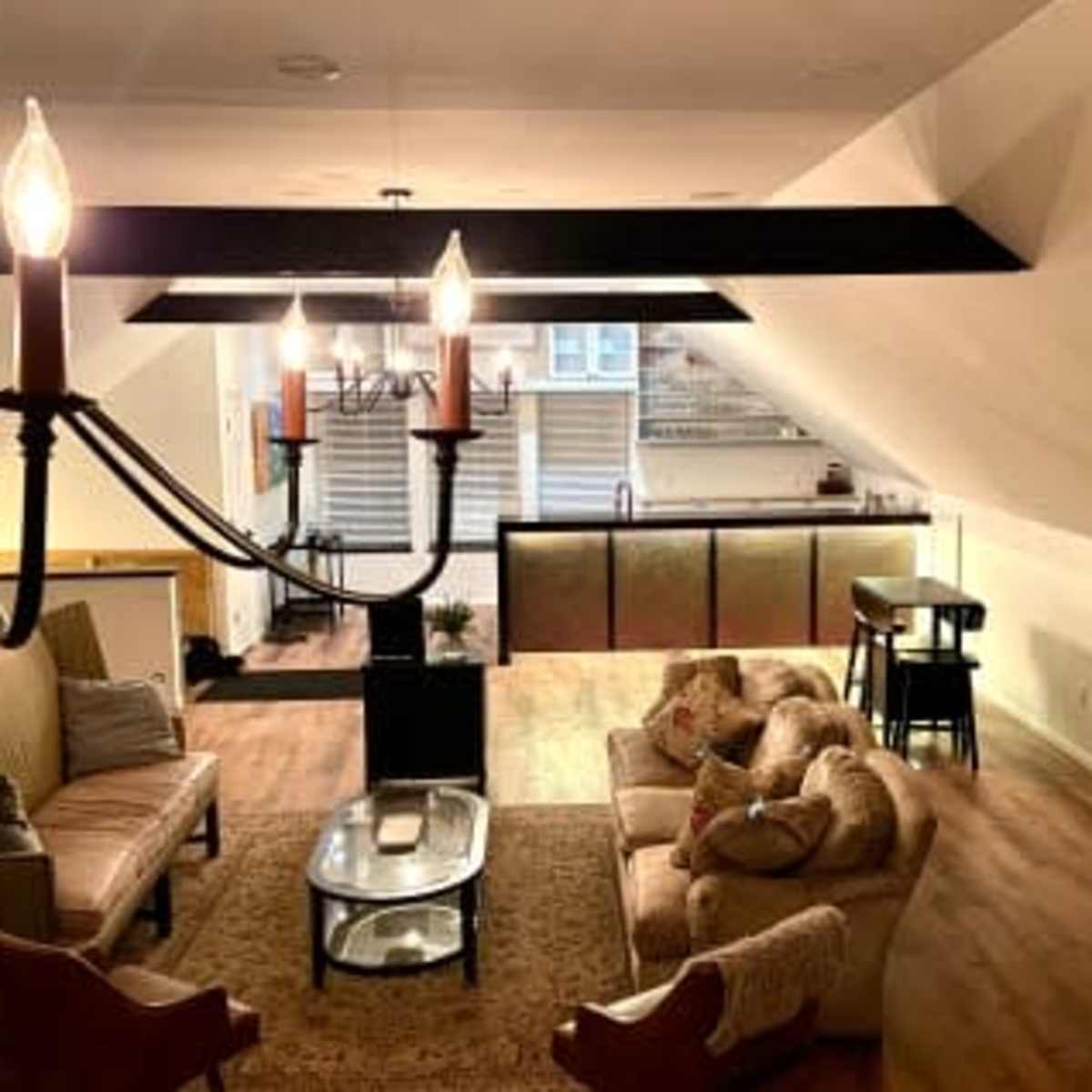 A spacious attic living area with a chandelier, a sectional sofa, a coffee table, and a kitchenette visible in the background.