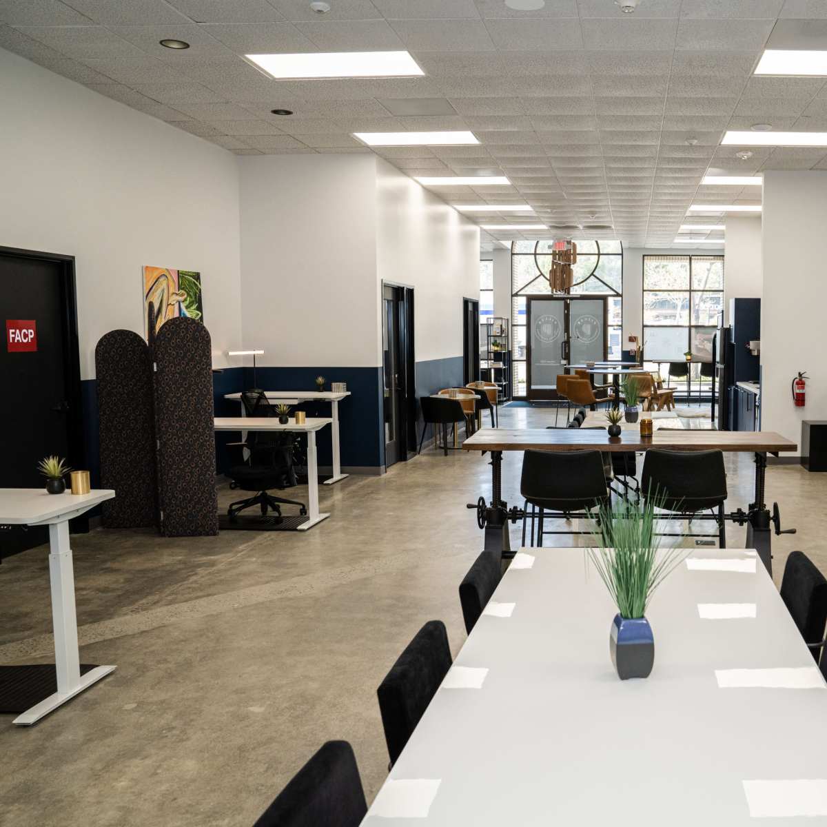 A modern office space with a large communal table, individual desks, and a glass-walled meeting room.