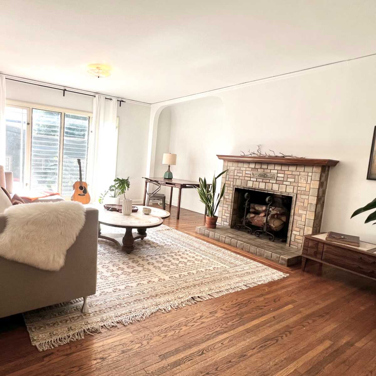 A cozy living room features a gray sofa, a wooden coffee table, a fireplace with a stone surround, and large windows allowing natural light.