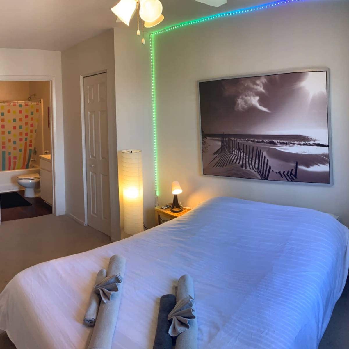 A spacious bedroom features a neatly made bed, a large landscape photograph on the wall, and a doorway leading to a brightly colored bathroom.