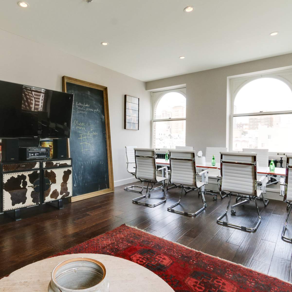 A modern office space features a large television, a wooden cabinet, a chalkboard wall, and a conference table surrounded by white chairs.