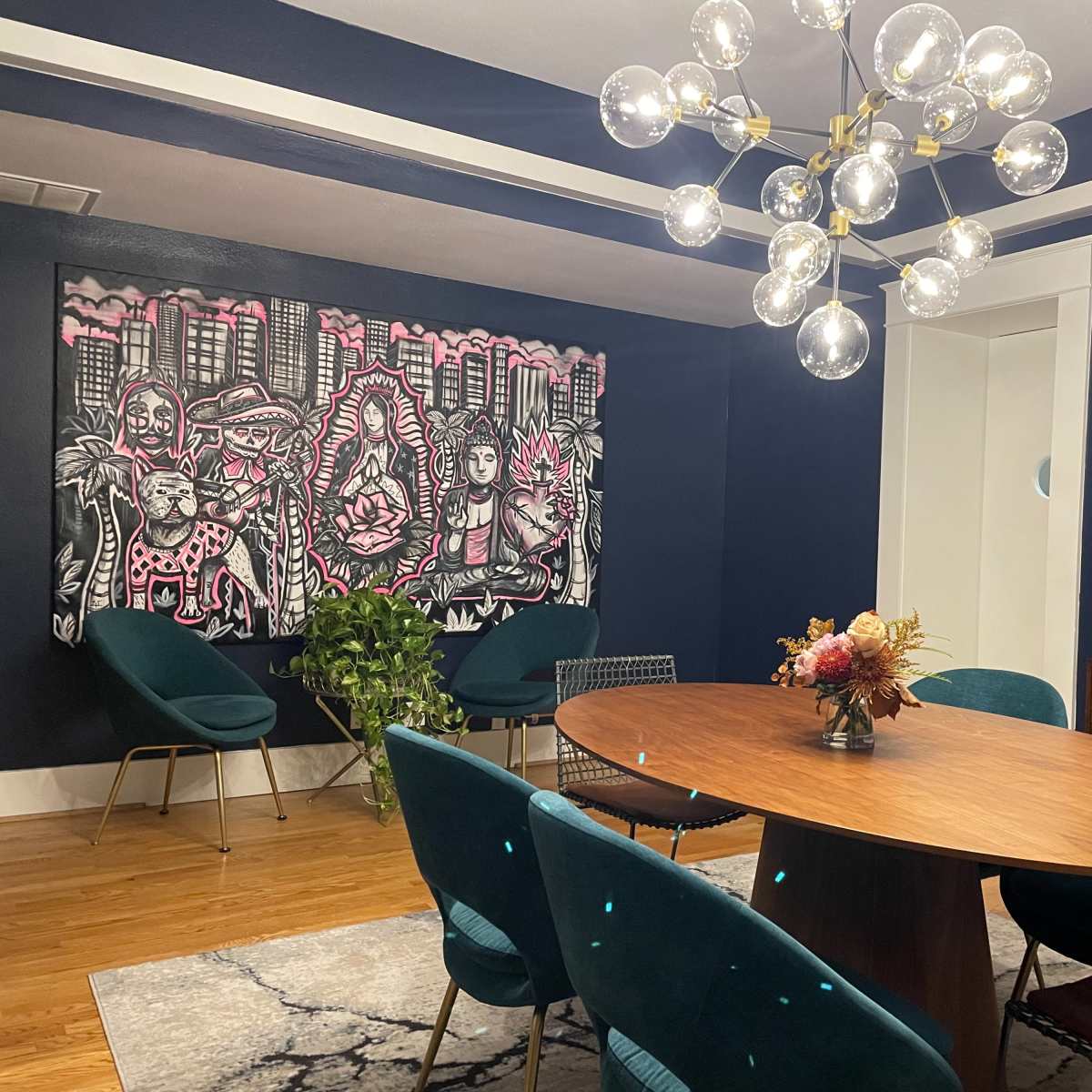A dining room features a round wooden table surrounded by teal chairs, with a large mural on the wall and a modern chandelier overhead.