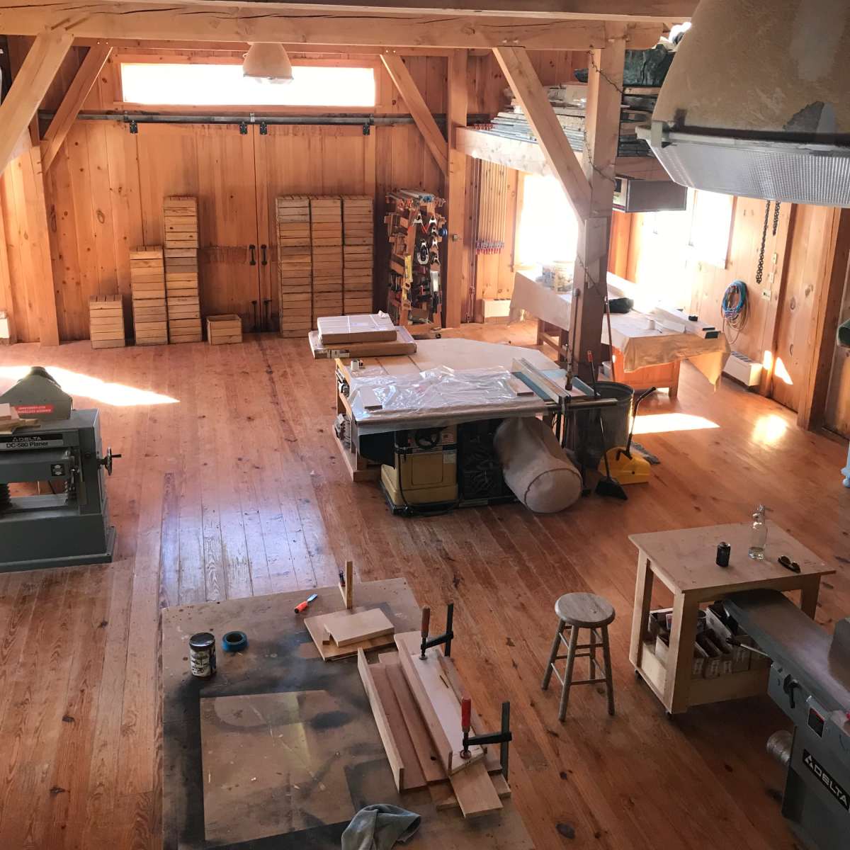 A spacious woodworking shop with wooden beams, tools, and workstations arranged neatly in a sunlit environment.