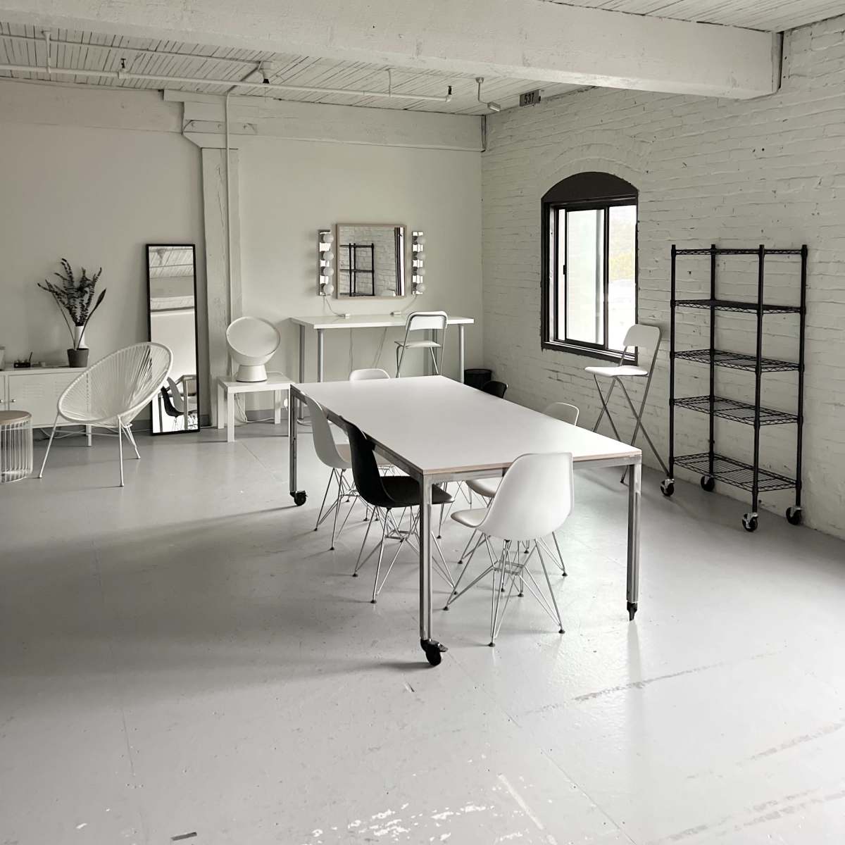 A spacious, well-lit room featuring a long table surrounded by chairs, a mirror on the wall, and a shelving unit.