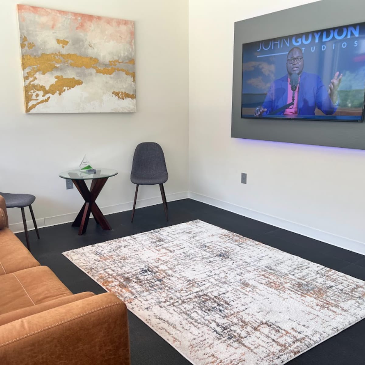 A modern sitting area with a brown sofa, a round wooden table, two chairs, a patterned rug, a wall-mounted television displaying a program, and artwork on the wall.