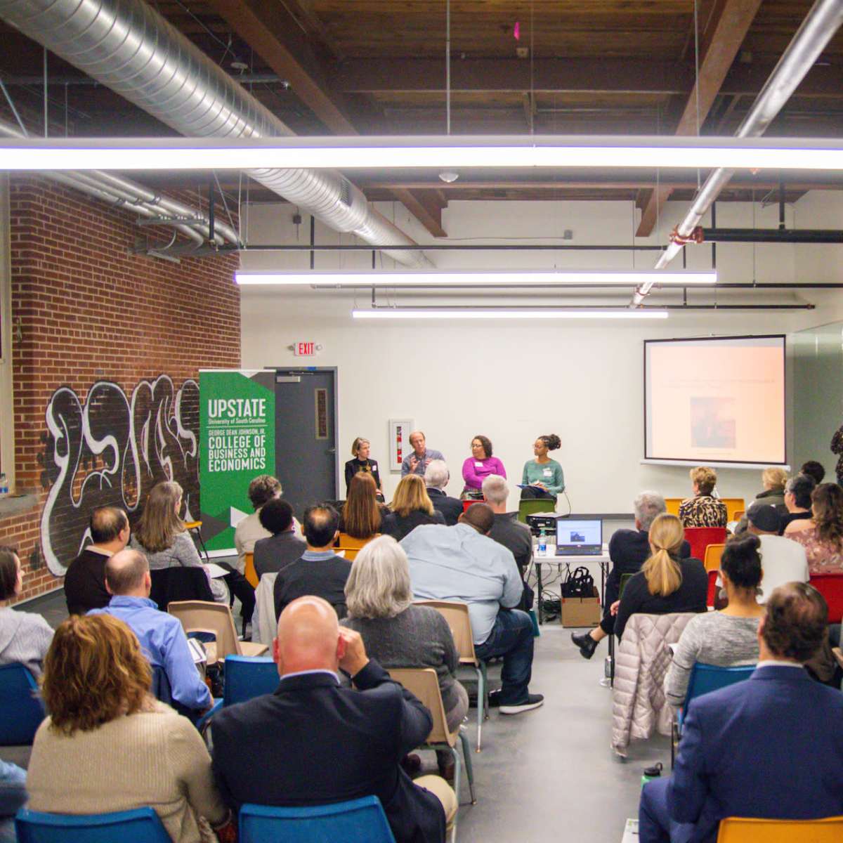 A diverse group of people attends a presentation in a spacious room with brick walls and modern lighting.