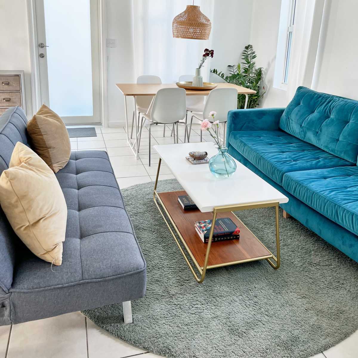 A bright living room featuring a gray sofa with pillows, a teal couch, a glass coffee table on a round green rug, and a dining area with a wooden table and white chairs.