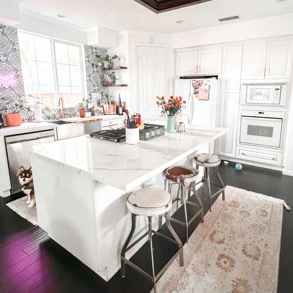 A modern kitchen with a large white island, three metal stools, and a variety of appliances and plants in the surrounding area.