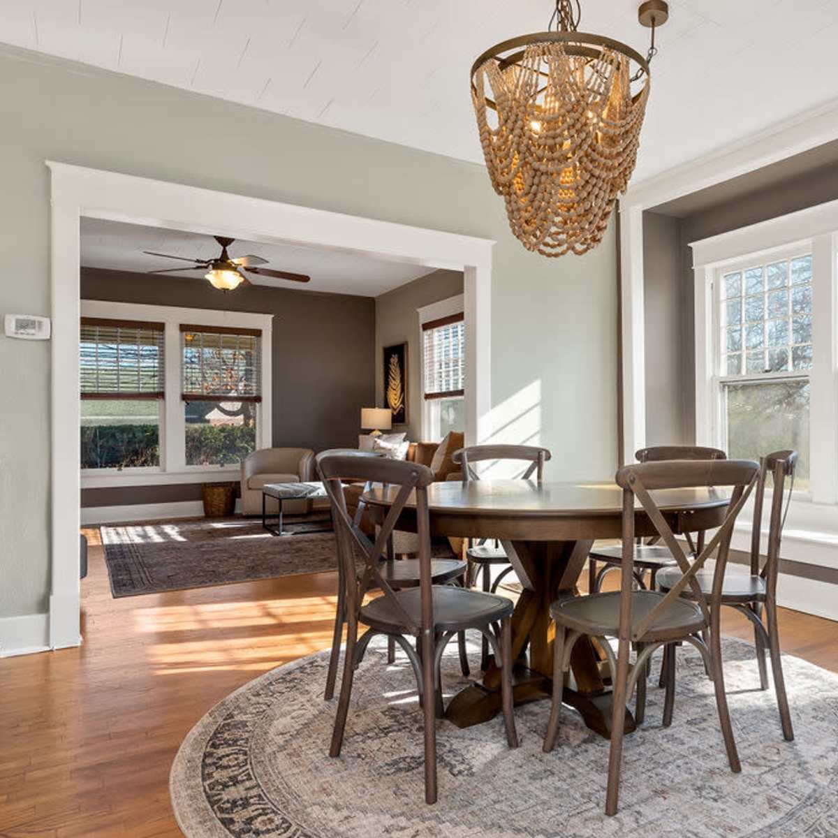 A dining area features a round table surrounded by chairs, illuminated by a chandelier, with large windows providing natural light.