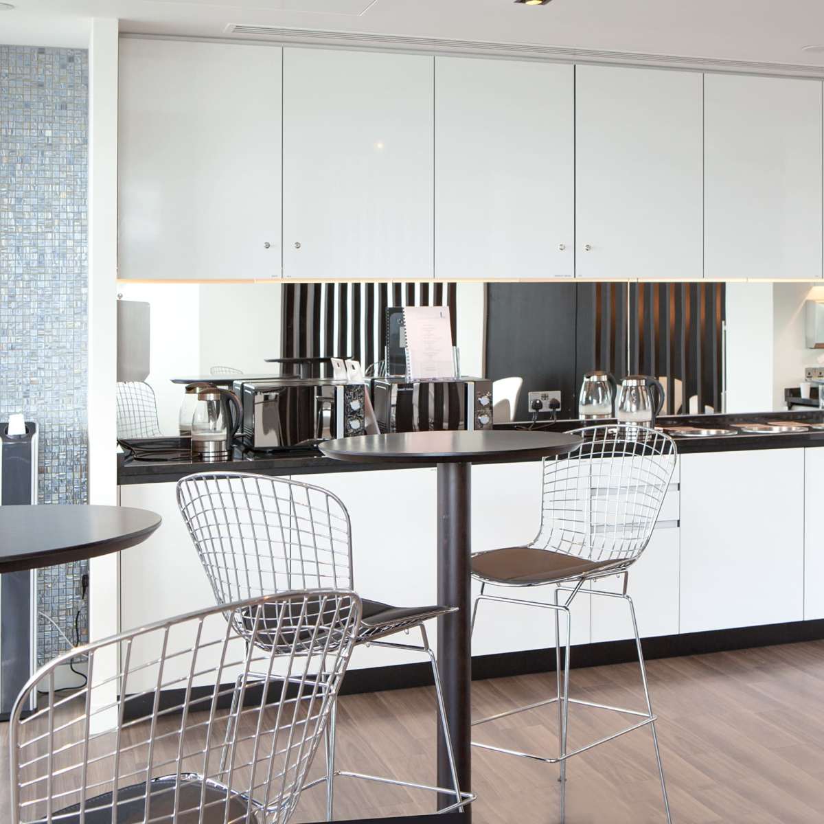 A modern kitchen with white cabinets, stainless steel appliances, and bar-height tables with wire chairs.