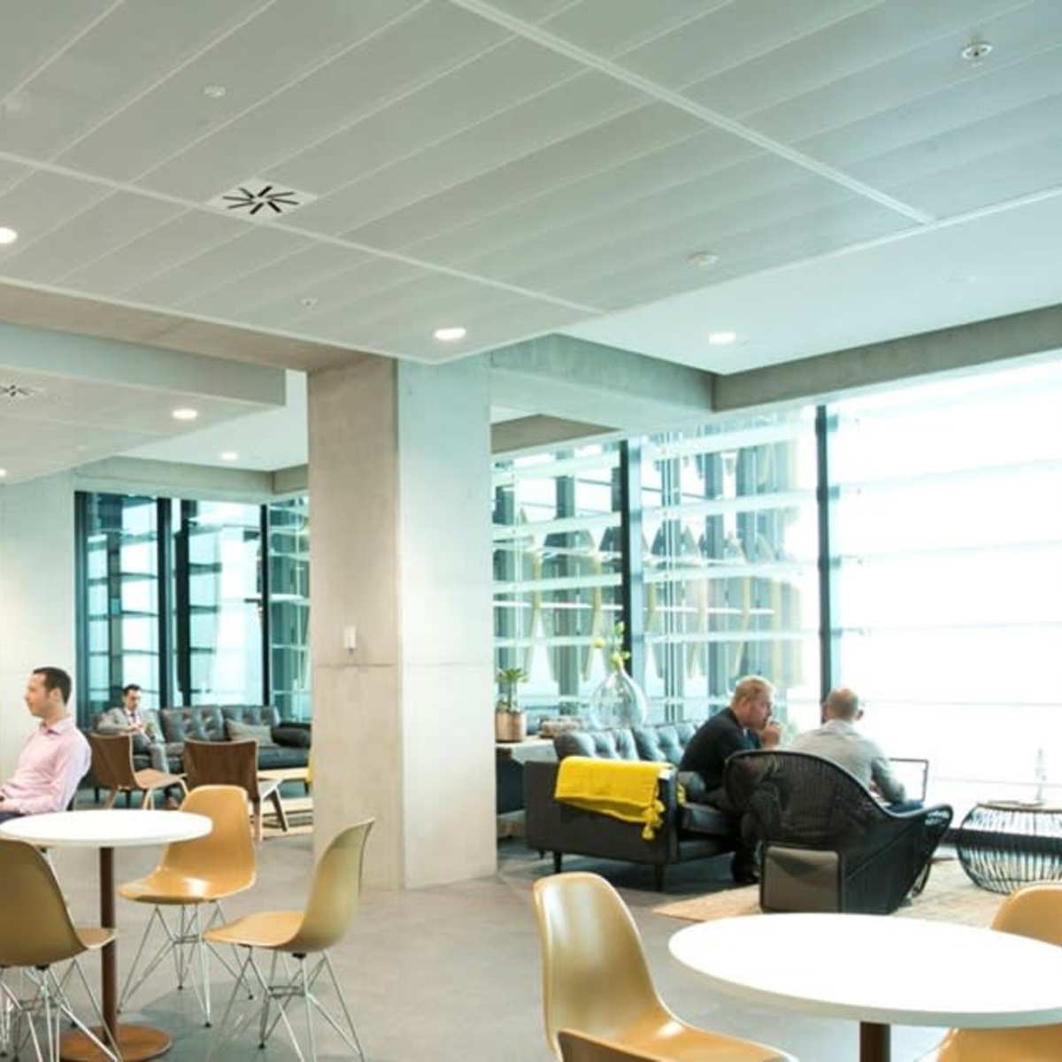 A modern lounge area with tables and chairs, featuring people seated and working, and large windows allowing natural light to enter.