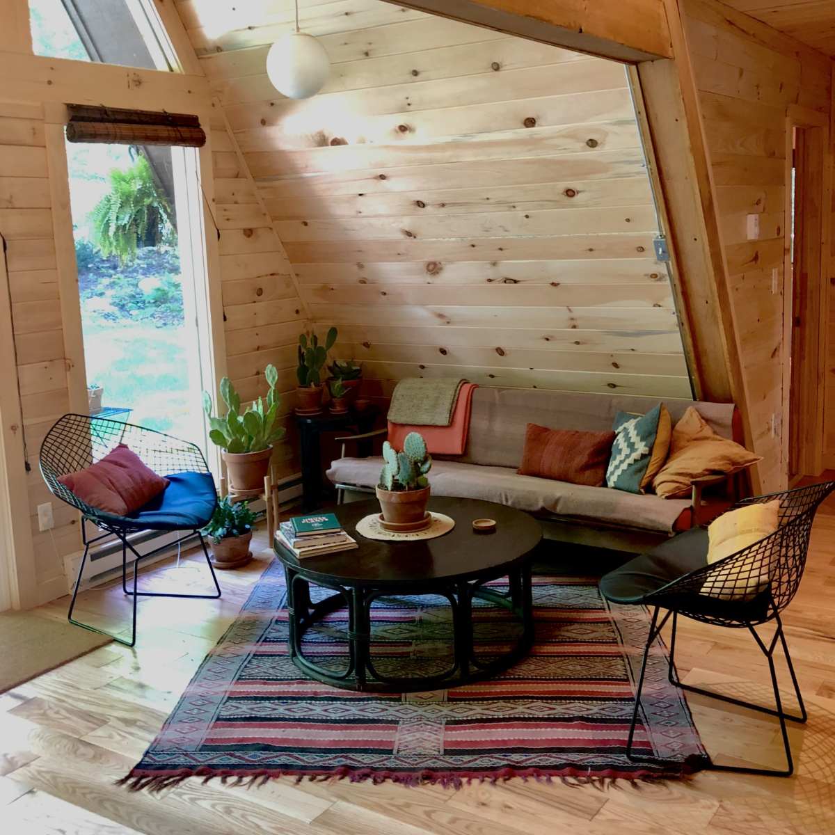 The interior features a wooden A-frame living room with a sofa, two wire chairs, a circular coffee table, and various potted plants, all arranged on a patterned area rug.
