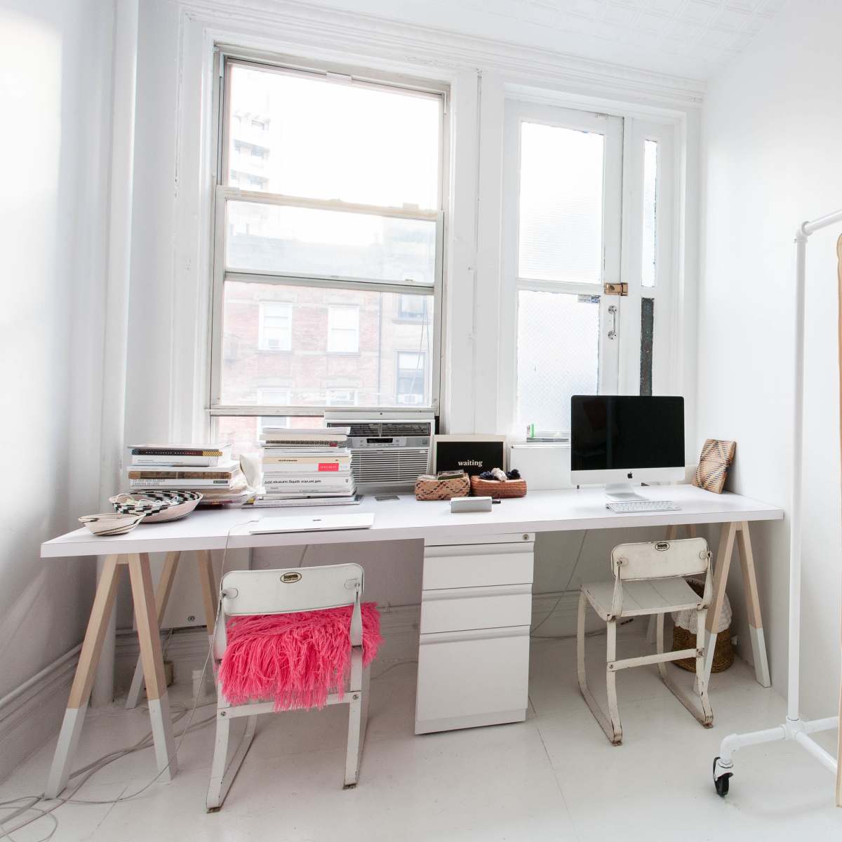 A minimalist office space featuring a white desk with a computer and books, alongside a clothing rack displaying several garments.