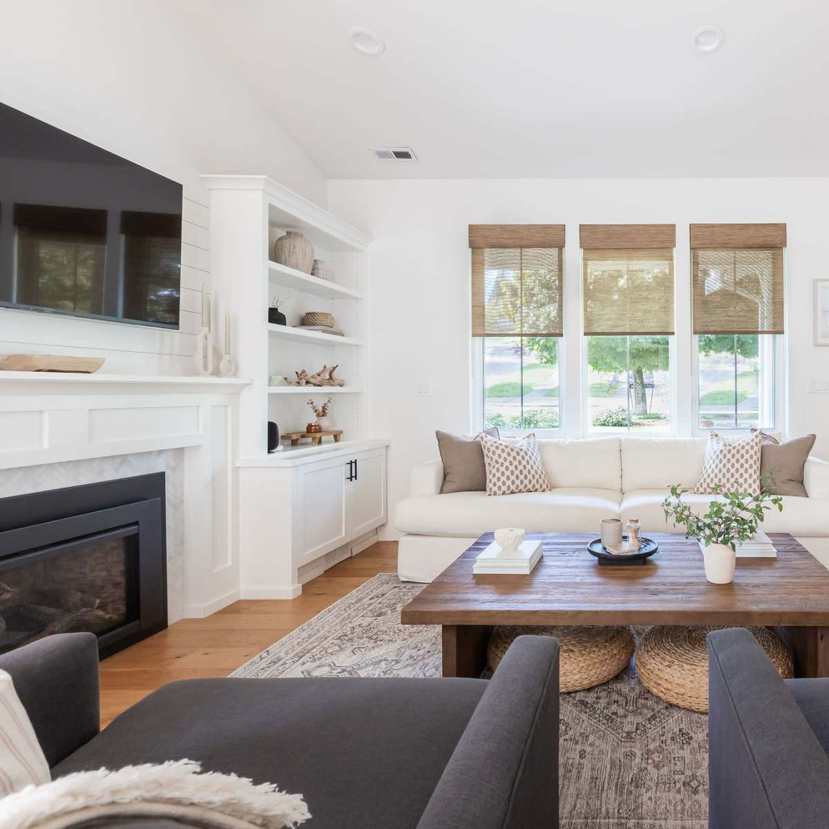 A modern living room features a white sofa, a wooden coffee table, and two gray chairs, with large windows allowing natural light and a view of greenery outside.