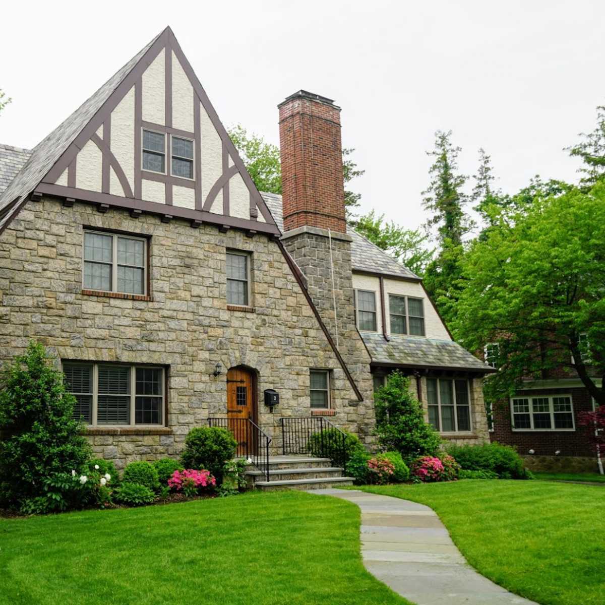A stone house with a steeply pitched roof and a central wooden door is surrounded by a well-maintained lawn and shrubs.
