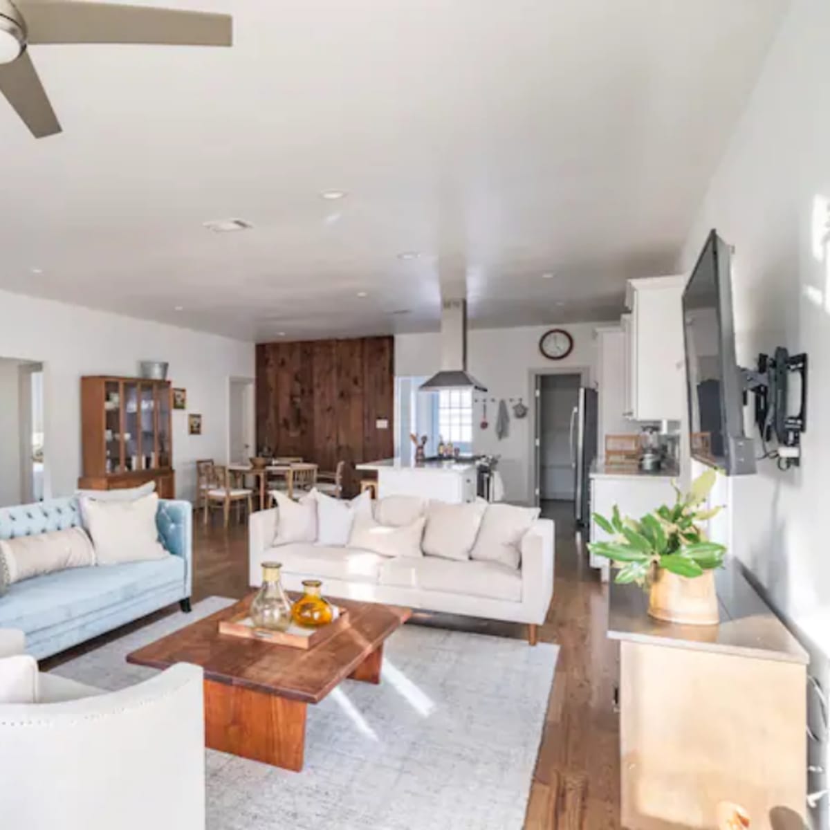 A spacious living room with a light-colored couch, a blue upholstered sofa, a wooden coffee table, and a kitchen area in the background.