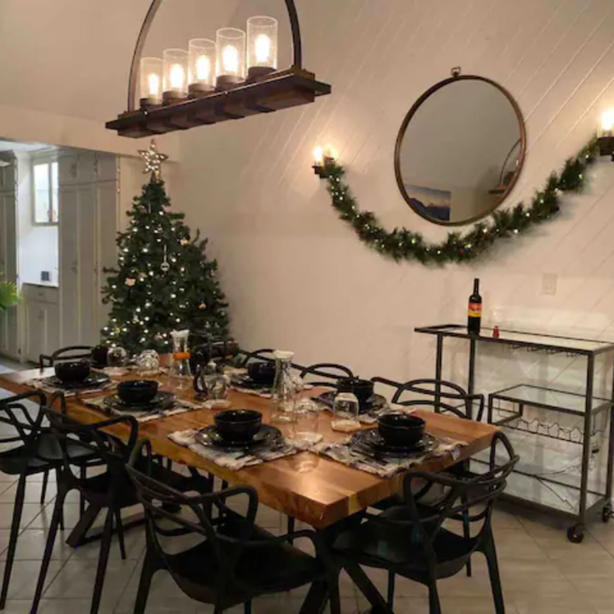 A dining area with a long wooden table set for a meal, surrounded by black chairs, a Christmas tree in the corner, and decorative lighting overhead.