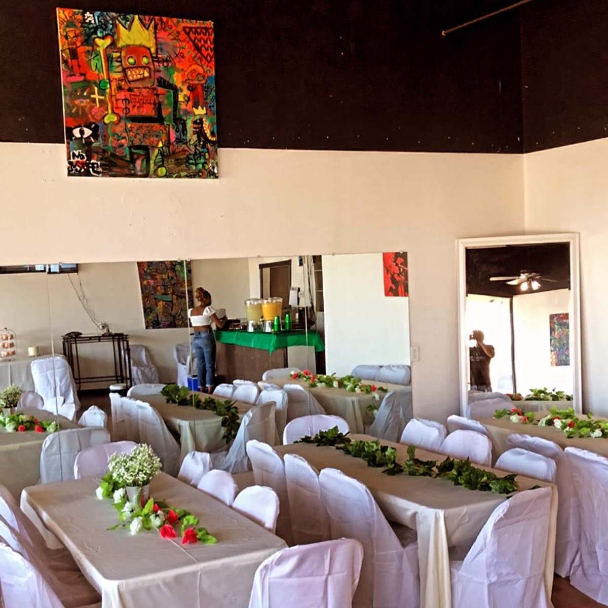 A dining area with multiple tables covered with white tablecloths and decorated with greenery is set up for an event.