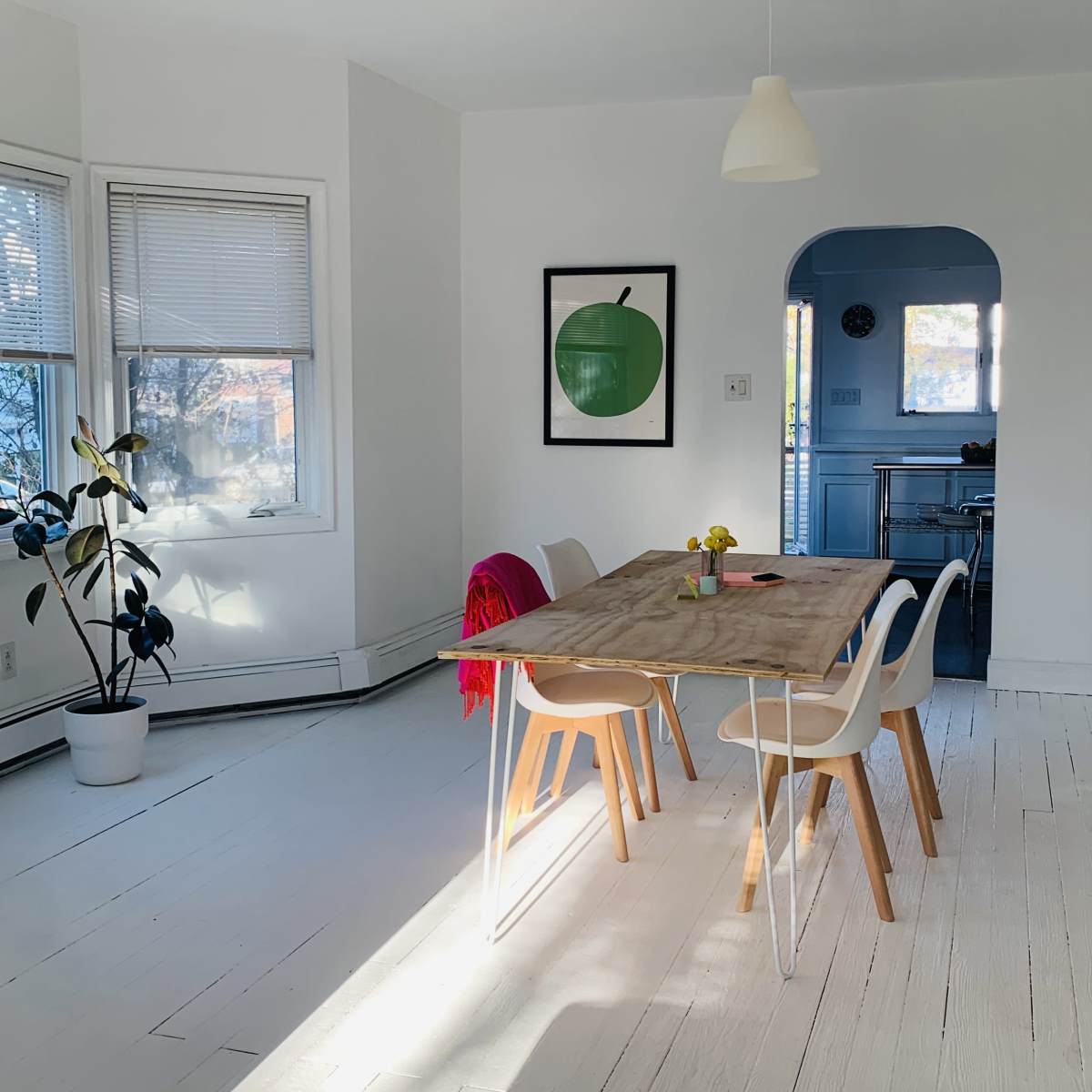 A bright dining area features a wooden table surrounded by white chairs, with a large green apple print on the wall and a potted plant in the corner.