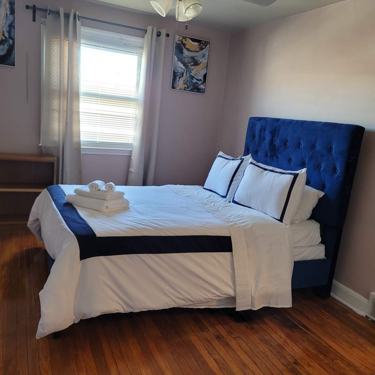 A well-organized bedroom featuring a blue upholstered bed with white linens and towels neatly arranged on it, accompanied by wooden flooring and minimal decor.