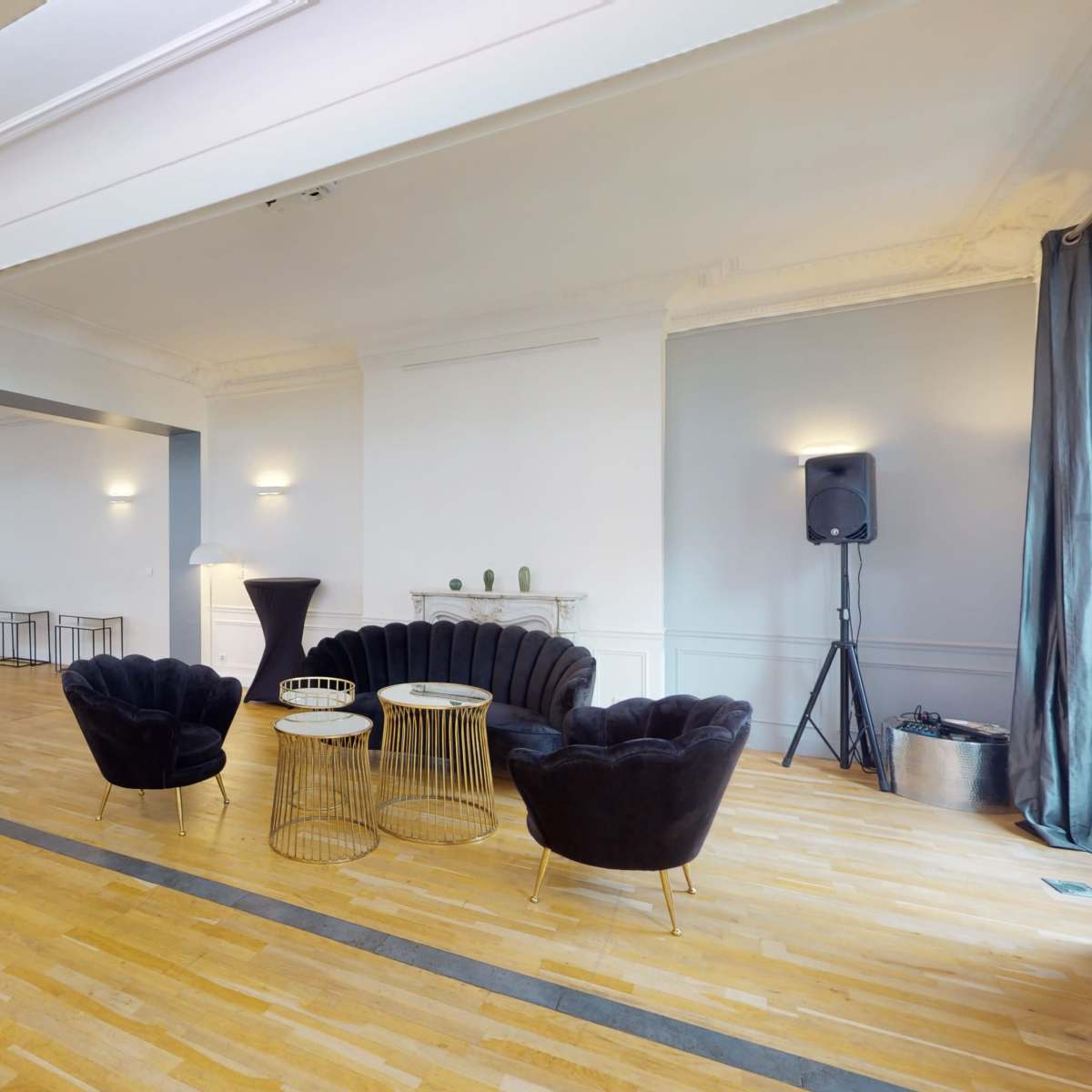 A spacious room featuring a set of black velvet chairs and small tables, with a speaker located in the corner and large windows letting in natural light.