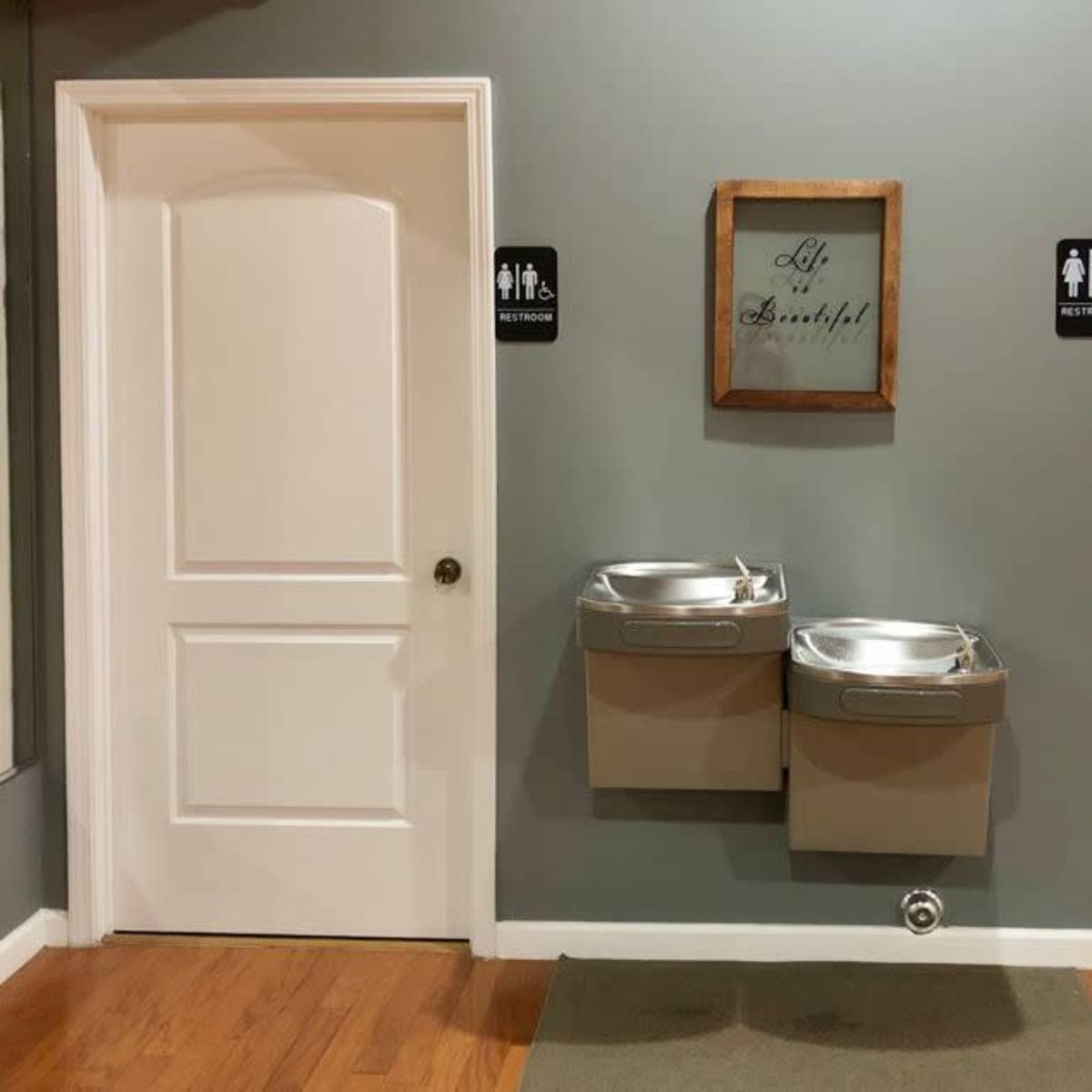 A hallway with two restroom doors on either side, a sign above reading "All Blessed," and two water fountains in the center.