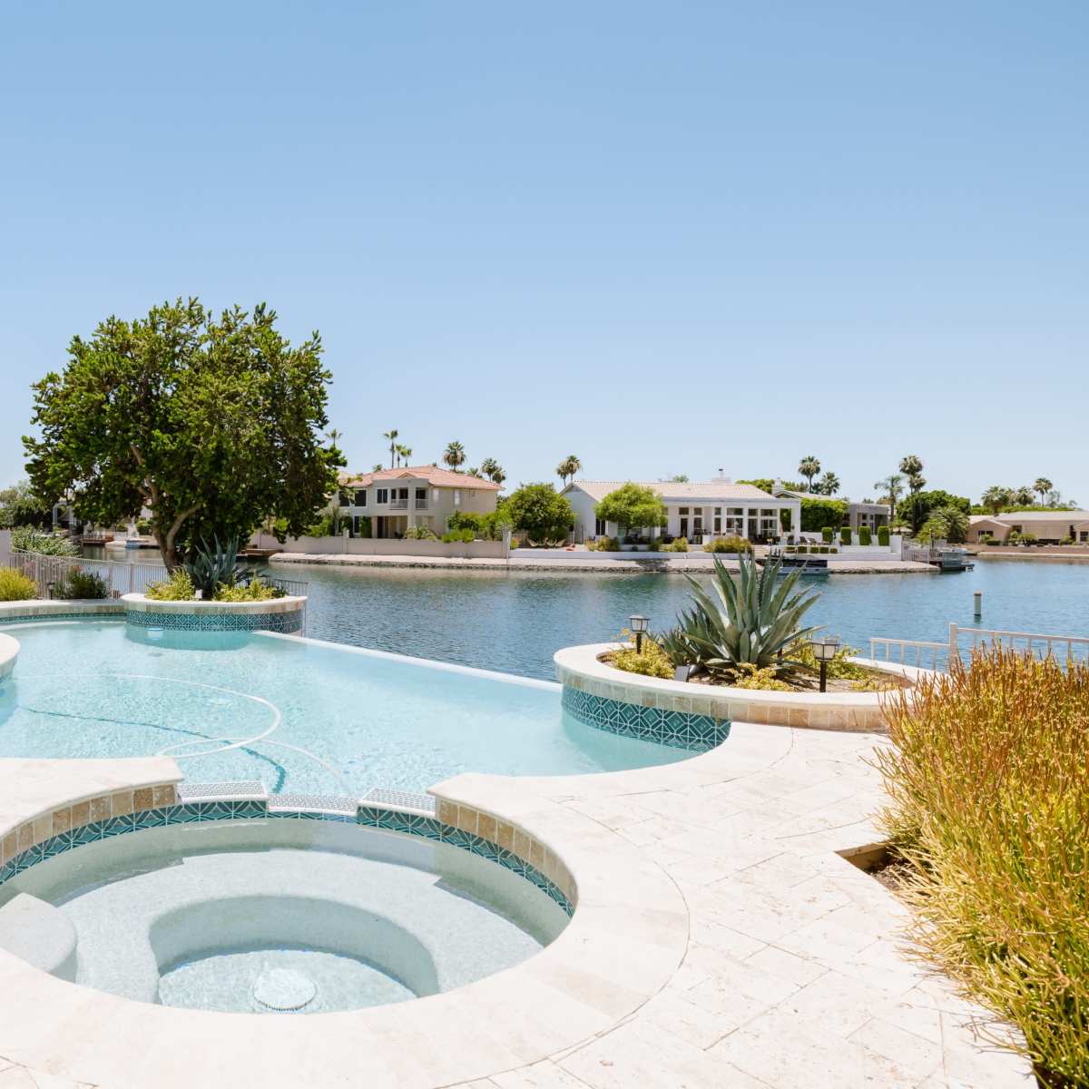A landscaped pool area with a spa overlooking a calm body of water and palm trees in the background.