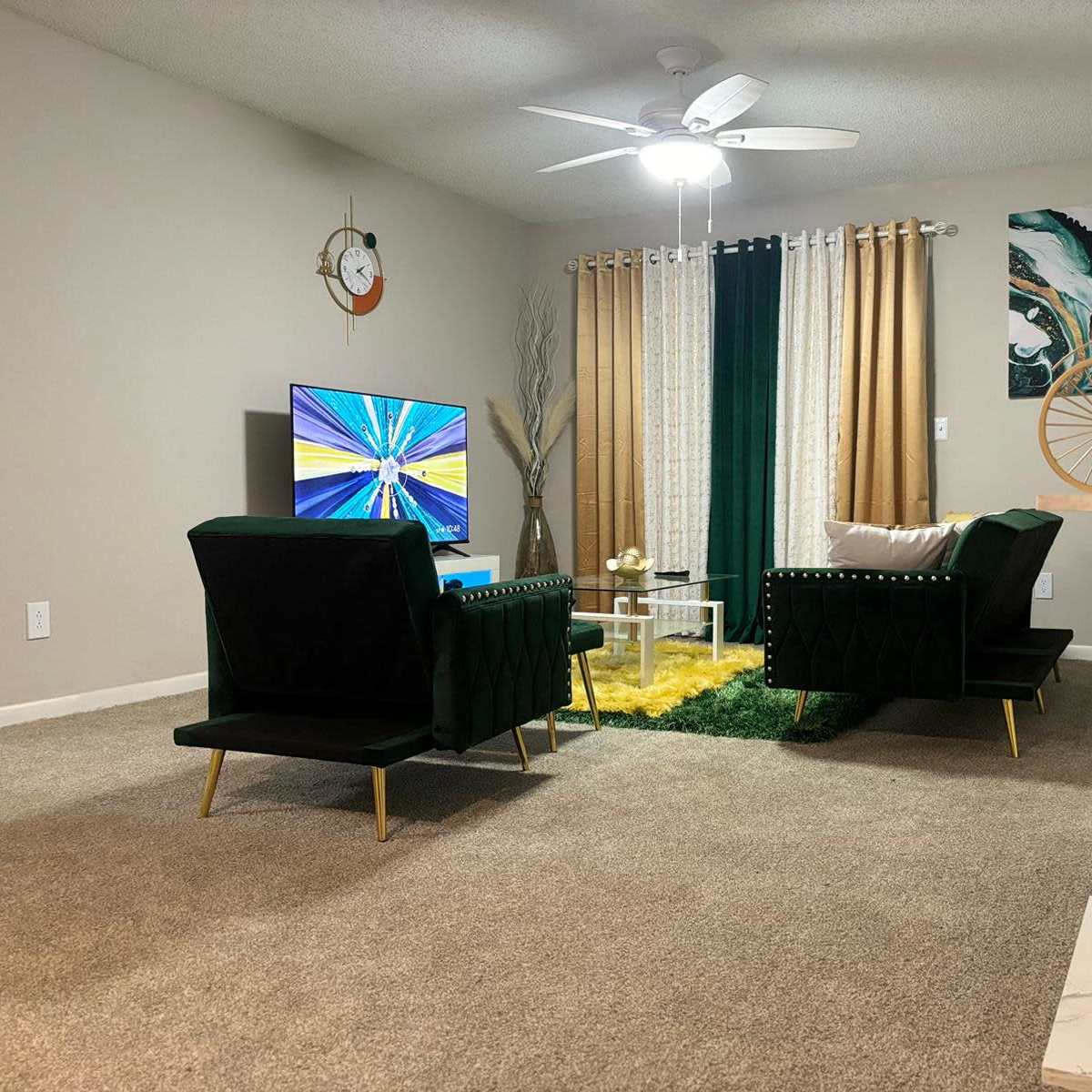 A living room features two green chairs, a television on a stand, and decorative elements, including a small table and a bicycle sculpture, with curtains and a rug adding texture to the space.