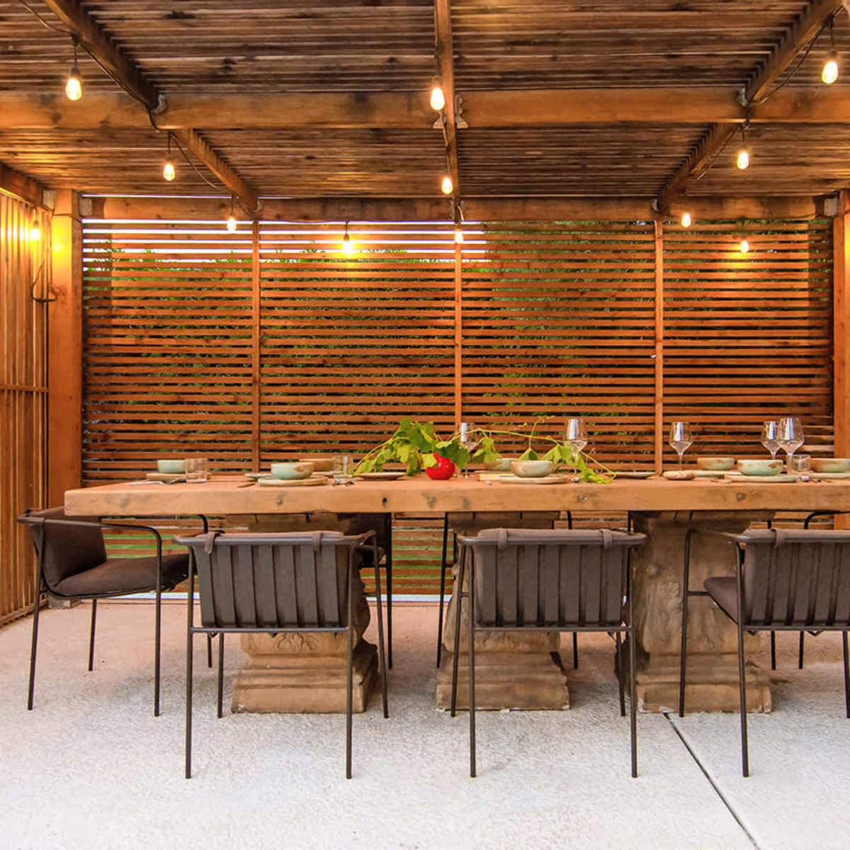 A wooden dining table with several chairs arranged under a pergola adorned with string lights, surrounded by wooden slatted walls.