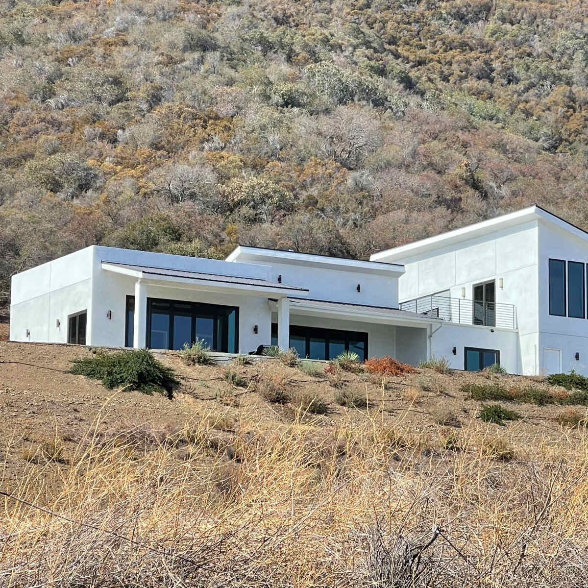 A modern, two-story white house is situated on a hillside with dry vegetation surrounding it.