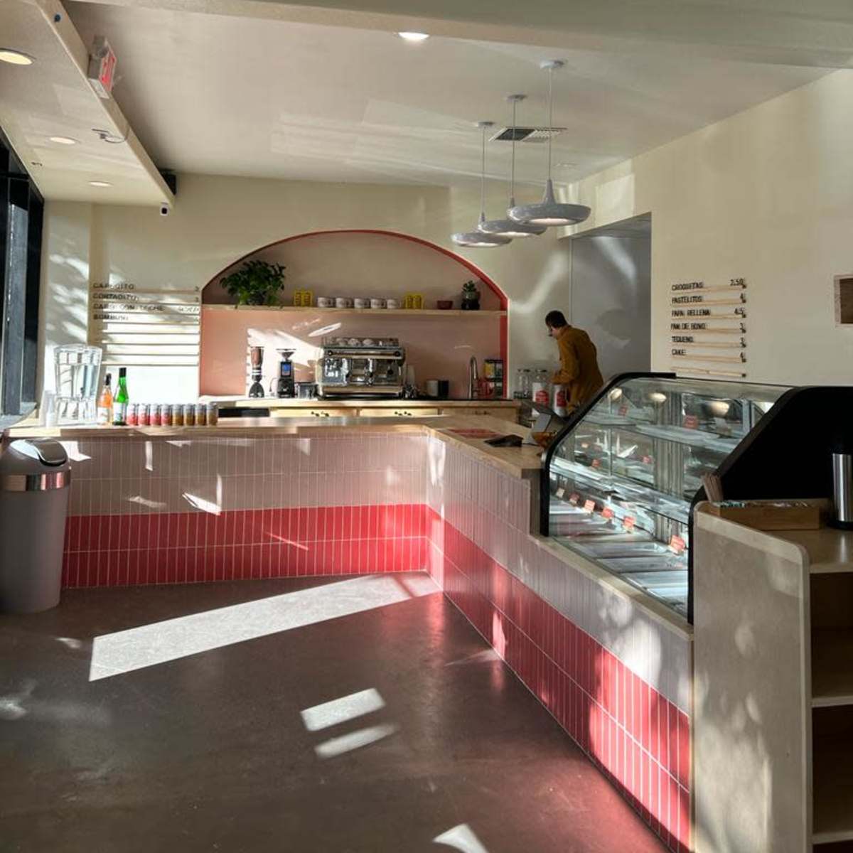 A modern cafe interior features a coffee station, display case, and a worker preparing items under bright overhead lights.