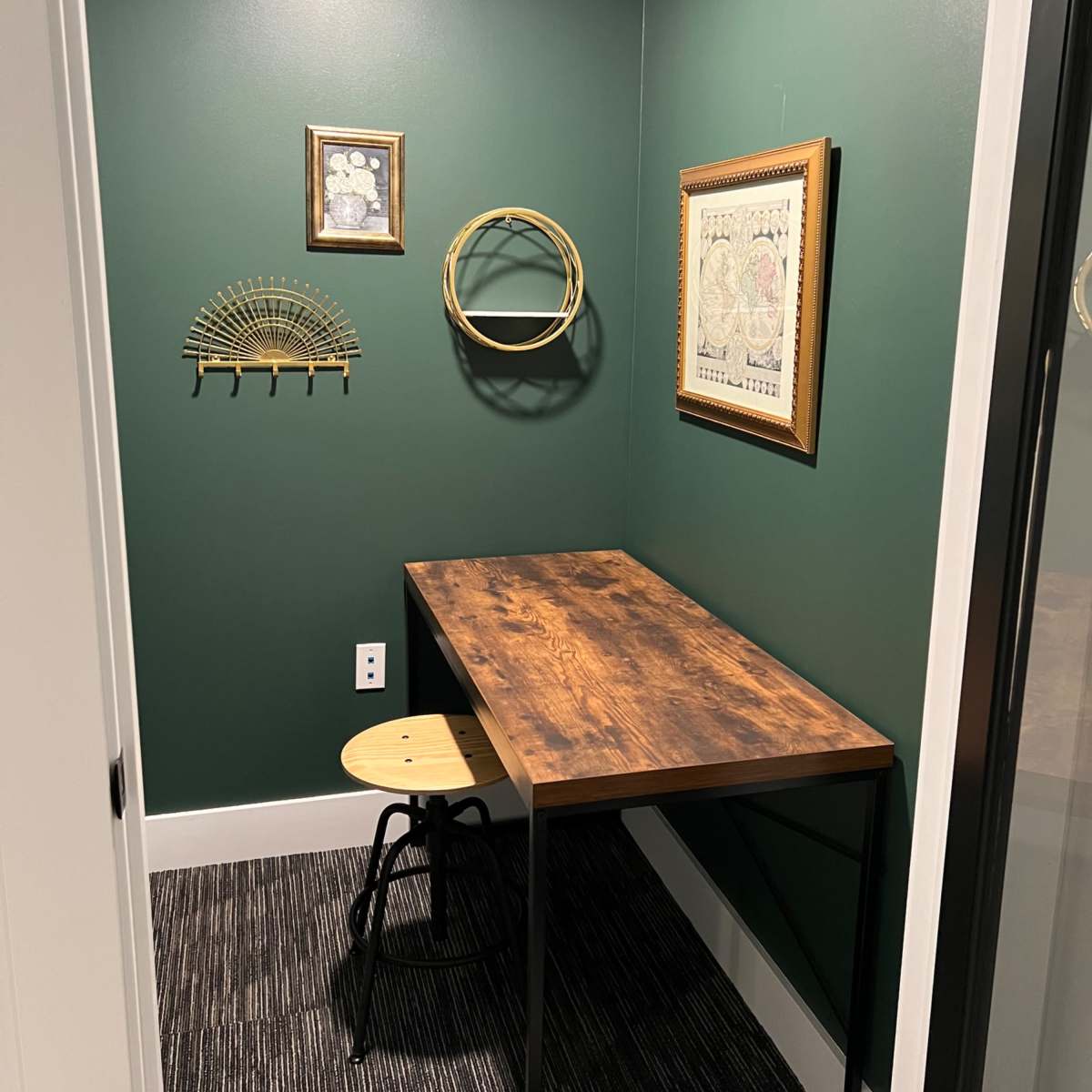 A small office space with a wooden desk and a stool, set against dark green walls adorned with framed wall art.