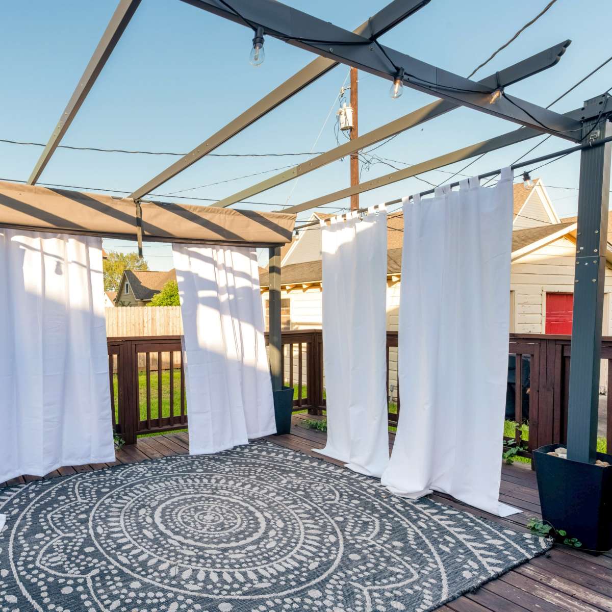 A covered wooden deck featuring white curtains hanging from a metal frame, with a round patterned rug on the floor.