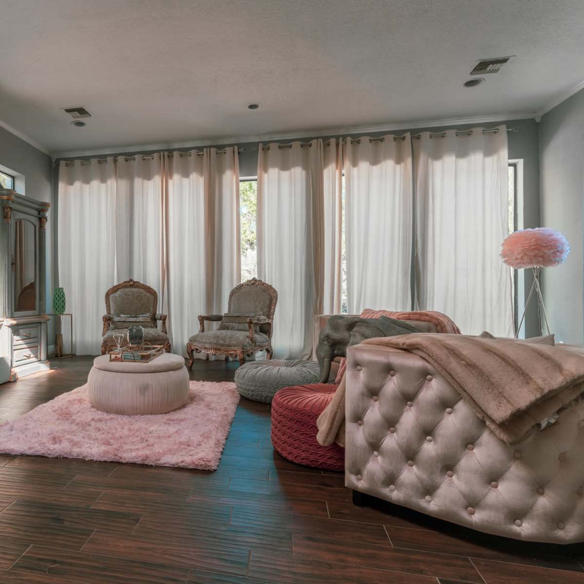 The room features elegant furniture, soft textiles, large windows with sheer curtains, and a cozy arrangement of seating areas.