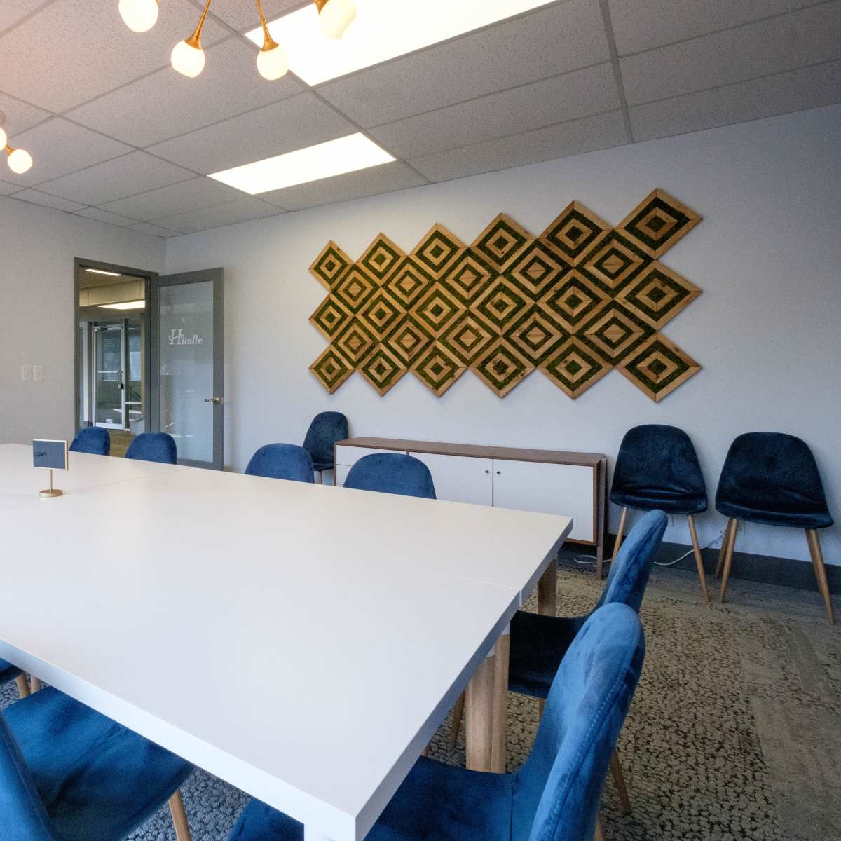 A spacious conference room features a large white table surrounded by blue chairs, with a decorative wall hanging in the background.