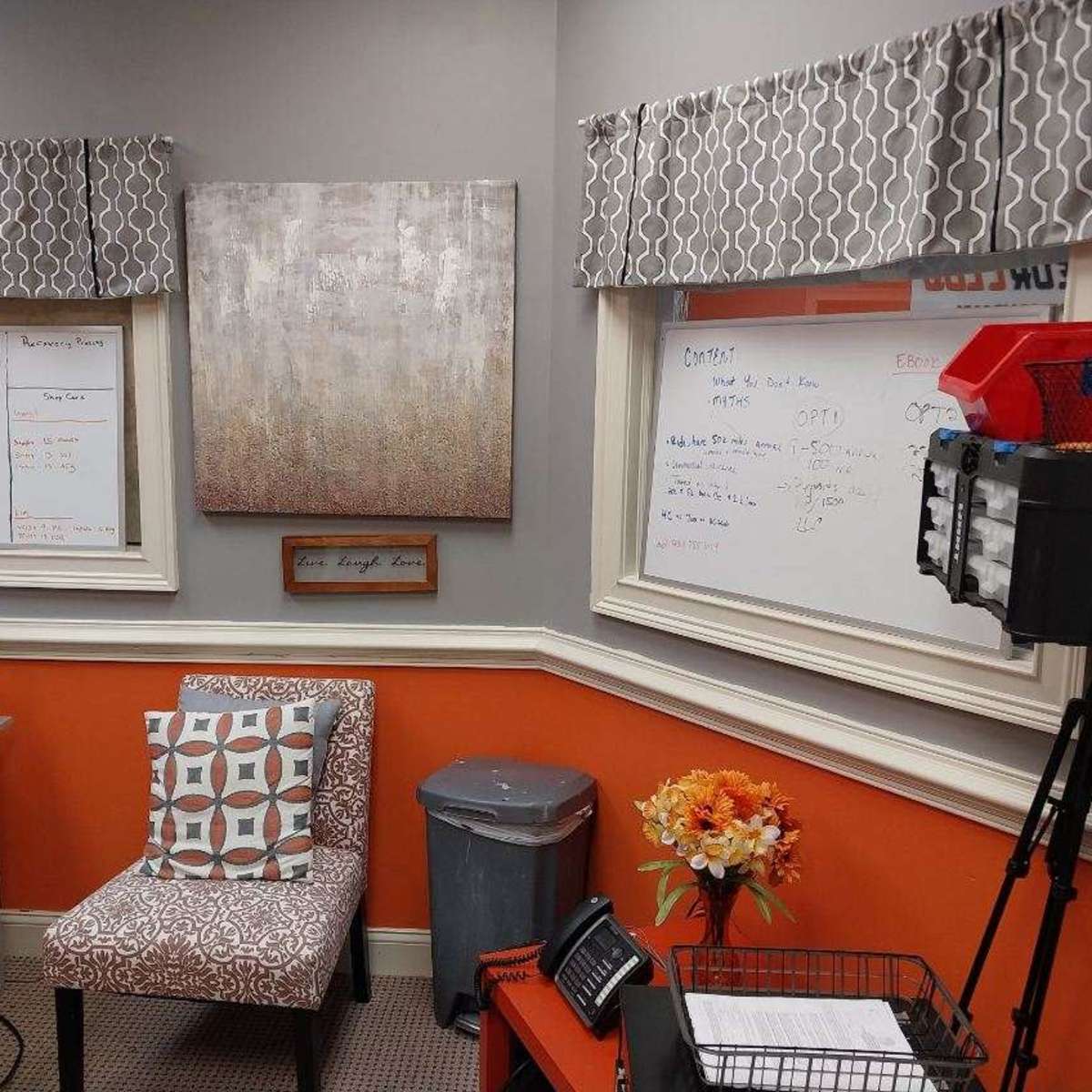 A small office with a gray chair, a whiteboard, and decorative curtains above two windows, featuring an orange wall and a trash can.