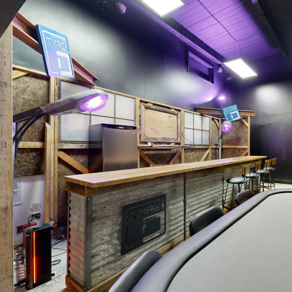 An interior room designed as a poker or gaming area, featuring a table with a grey felt surface, a bar with high stools, and purple lighting.
