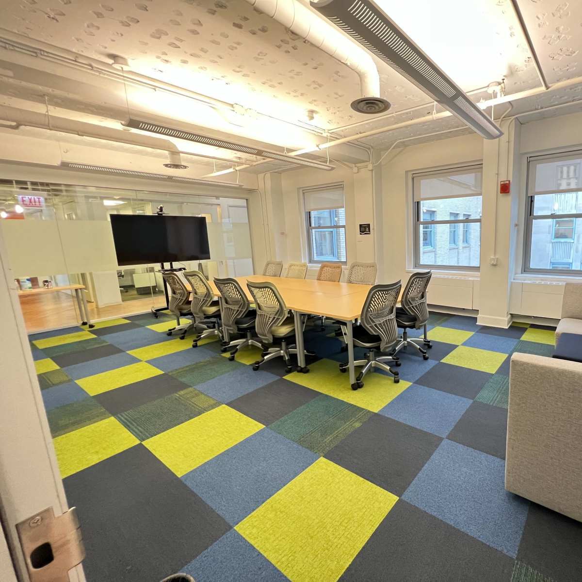 A conference room featuring a large table surrounded by office chairs, a television screen on the wall, and a gray sofa in the corner, with a multicolored carpet and windows providing natural light.