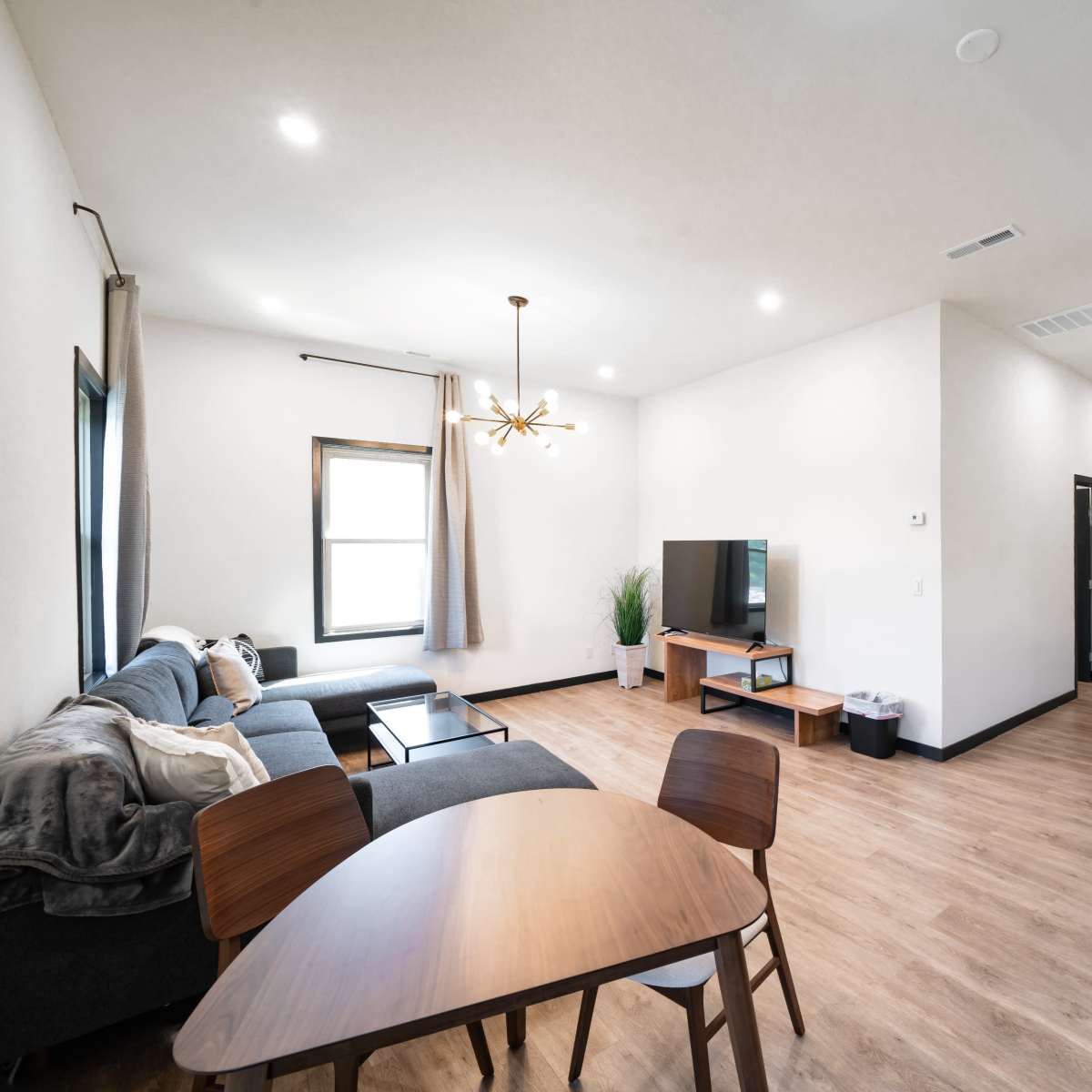 A modern, open living space featuring a gray sectional sofa, a wooden dining table, a television, and a kitchenette in a well-lit environment.