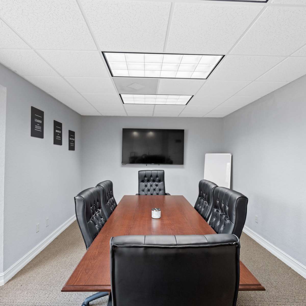 A modern conference room features a long wooden table surrounded by black leather chairs, with a television mounted on the wall and windows letting in natural light.
