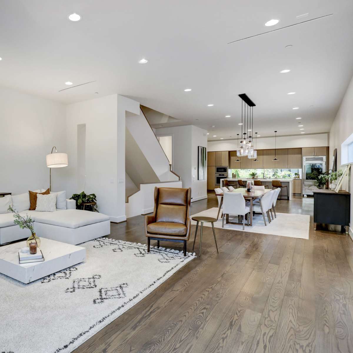 A modern living room with a white sectional sofa, a round mirror, and wooden flooring, adjacent to a dining area and kitchen.