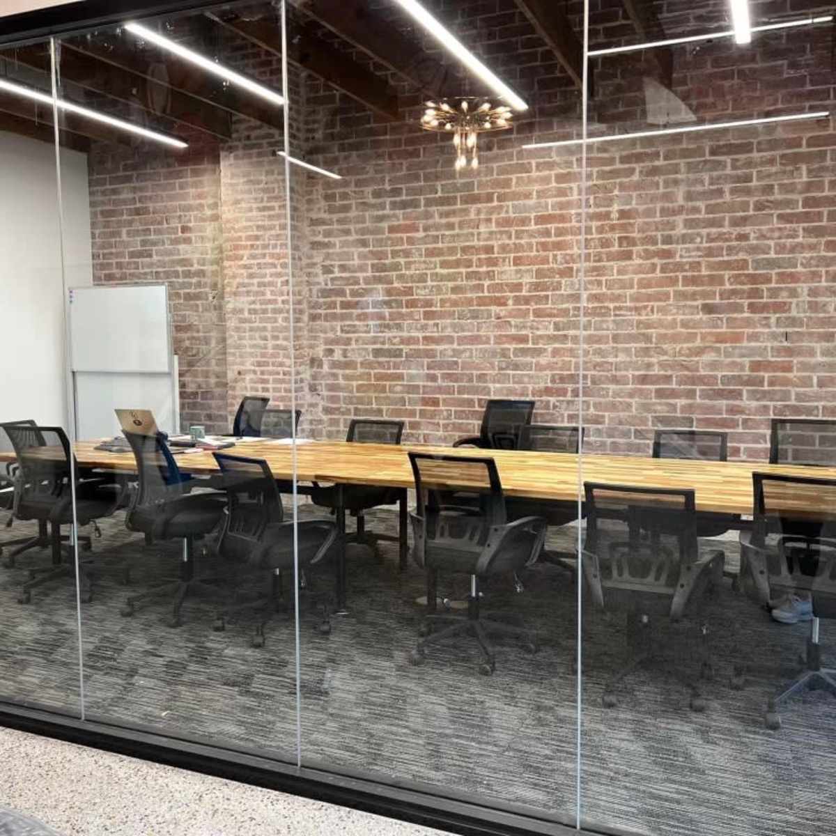 A modern conference room with a large wooden table surrounded by black office chairs, all enclosed by glass walls and featuring exposed brick and contemporary lighting.