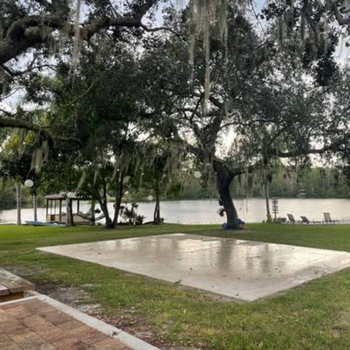 A concrete slab is positioned on a grassy area near a river, with a large tree draped in Spanish moss and a few lounge chairs visible in the background.