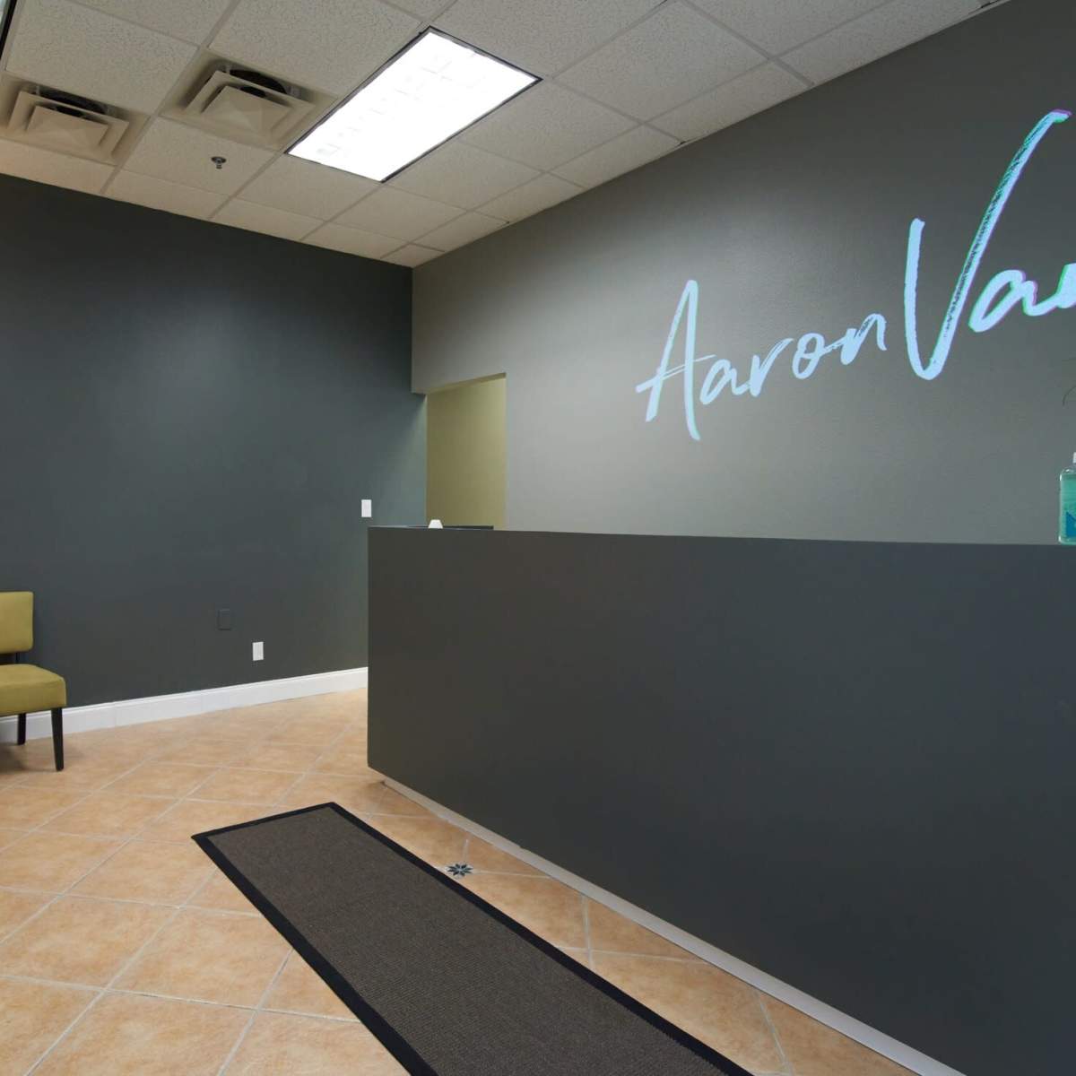 A modern reception area with a desk, two green chairs, and a decorative ladder shelf against a gray wall featuring the name "Aaron Van."