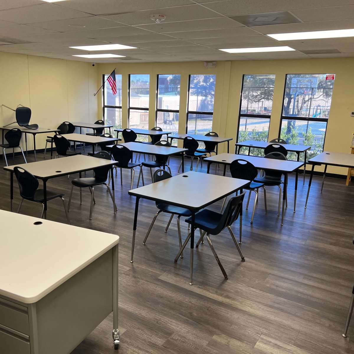 A classroom is arranged with multiple tables and chairs, featuring windows that overlook an outdoor area, and a desk positioned in the foreground.