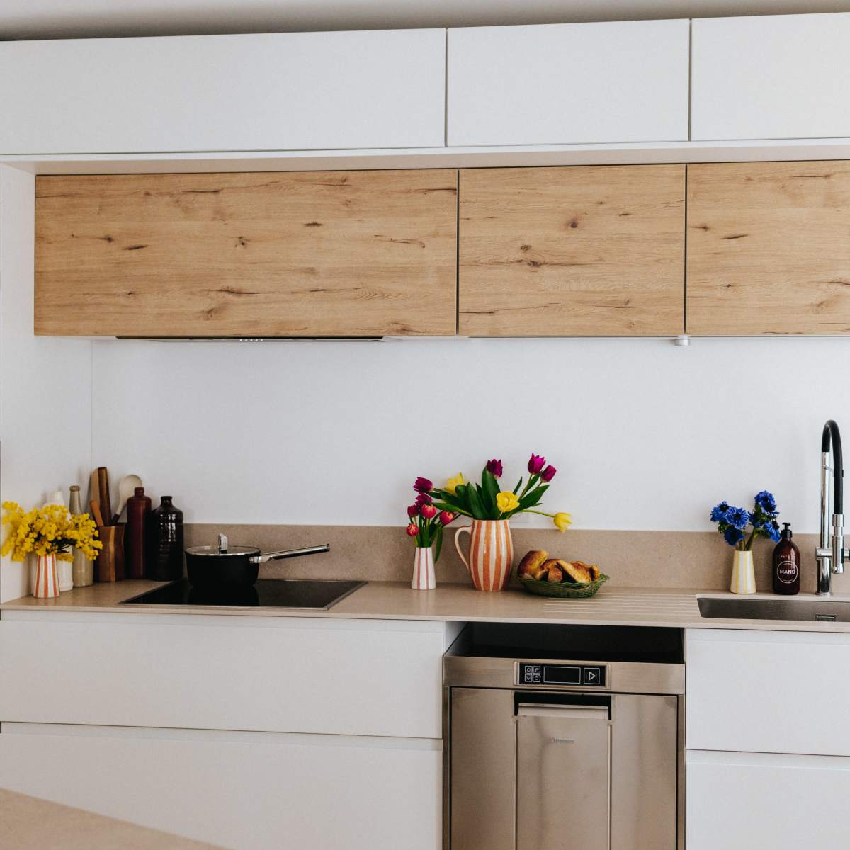 A modern kitchen features wooden cabinets, a stainless steel dishwasher, a sink, and vases of fresh flowers on the countertop.
