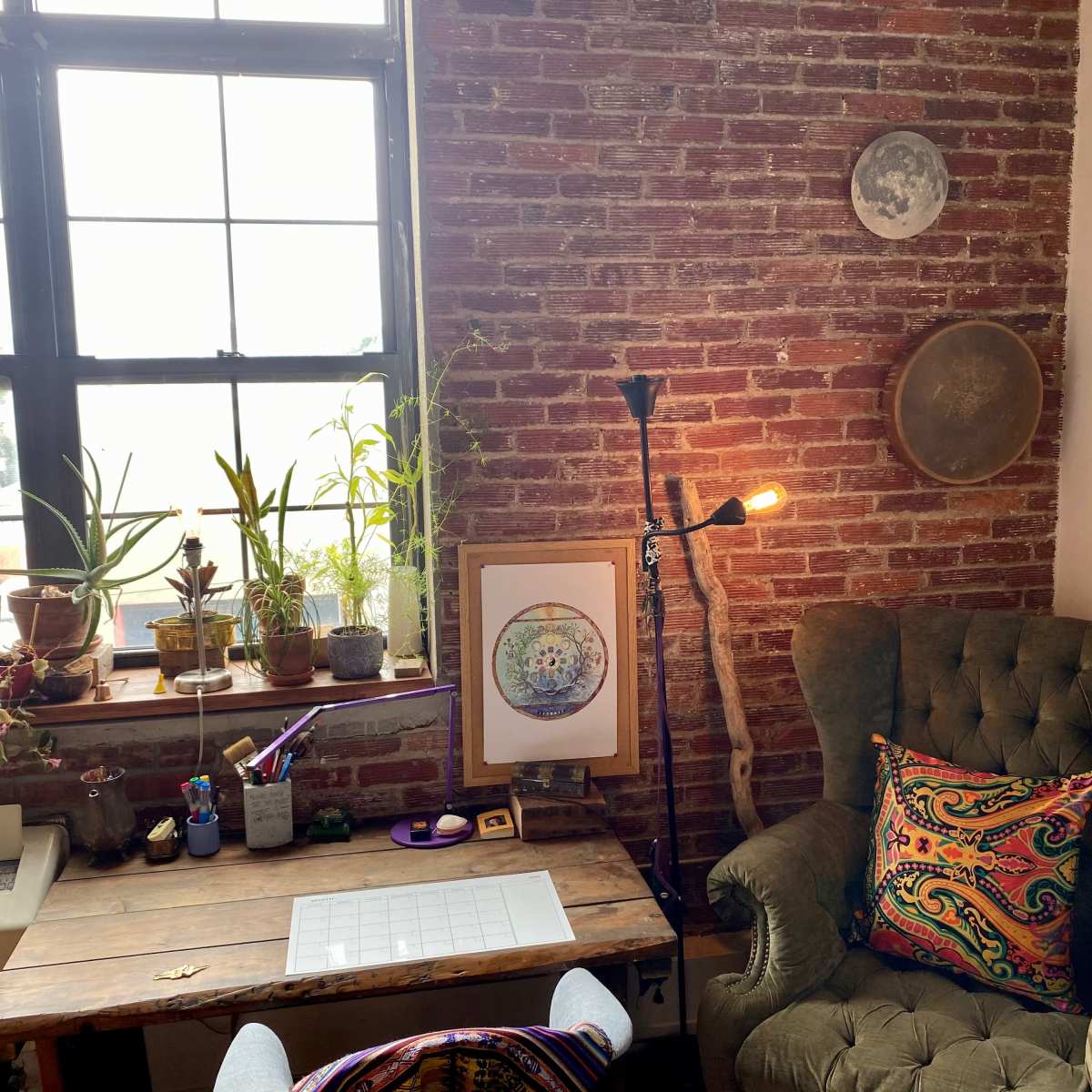A cozy home office with a vintage green armchair, a wooden desk, various plants on a windowsill, and warm lighting from a lamp.