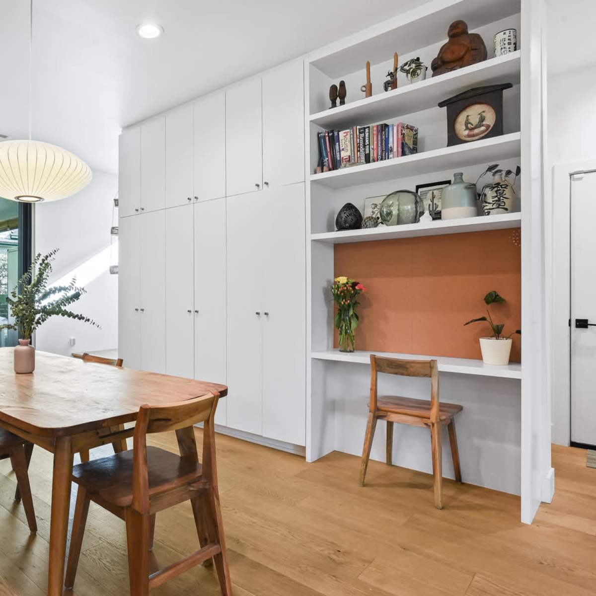 A dining area features a wooden table and chairs, with a built-in shelving unit displaying decorative items, and large windows that open to an outdoor space.