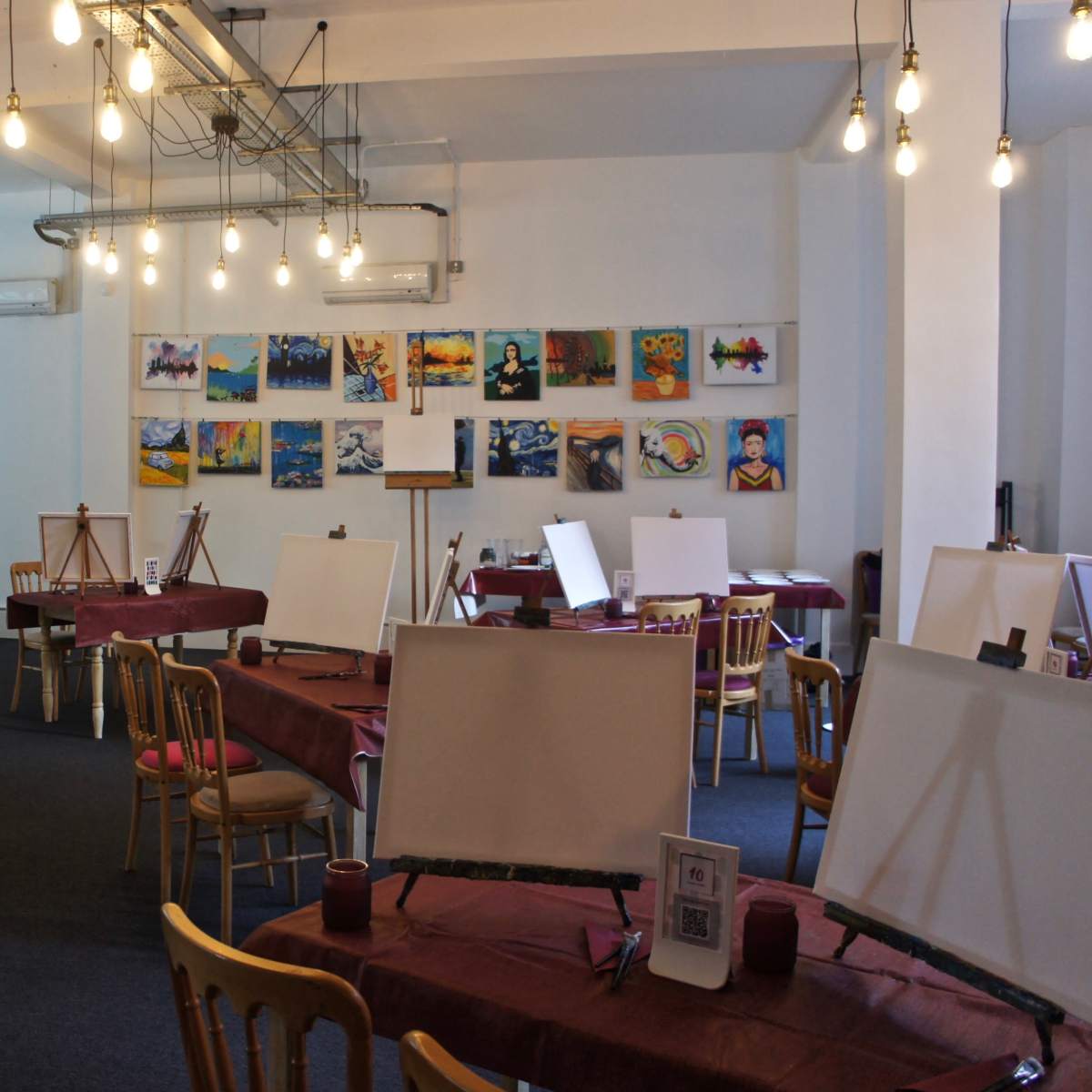 A spacious art studio with several empty easels arranged around tables covered in maroon cloths, illuminated by hanging light bulbs.