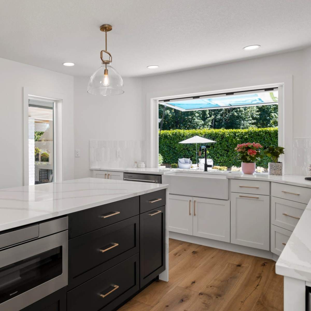 A modern kitchen with a large island, featuring a black countertop and stainless steel appliances, and a window that opens to an outdoor area.